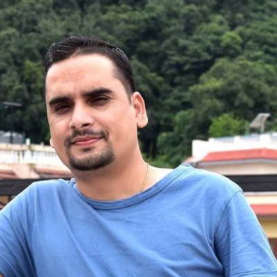 Aman Ghimire in a blue T-shirt, with rooftops and a forested hillside behind him.