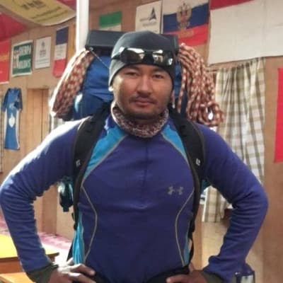 Buddhi Lama with his hands on his hips, in a blue base layer and a knit hat, with a pack and coiled climbing rope on his back, in a room hung with flags.