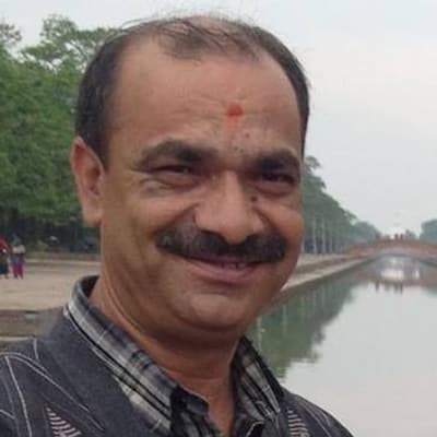 Kumoda Adhikari smiling, in a checked shirt, beside a long pool of water lined with trees.