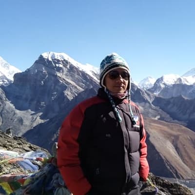 Partap Gurung in a red and black down jacket, a knit hat and sunglasses, on a rocky high point with prayer flags and snow peaks behind.