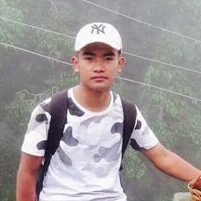Prakash Ghale in a white cap and a patterned T-shirt, wearing a backpack, with misty trees behind him.