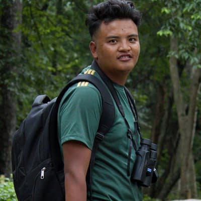 Samyam Rumba in a green T-shirt, with a black backpack and binoculars, looking over his shoulder in a forest.